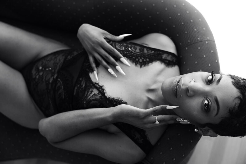 Black and white boudoir image of a woman with short hair and manicured nails, lying on a polka-dotted couch in San Francisco. Wearing a black lace lingerie bodysuit, she caresses her face and rests her hand on her breast, displaying a gaze that is both graceful and fiercely elegant