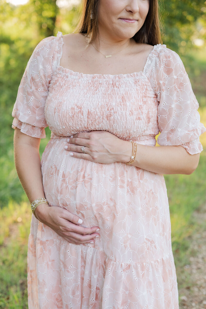 Woman in pink dress holding her belly with greenery background during Maternity Photography Session