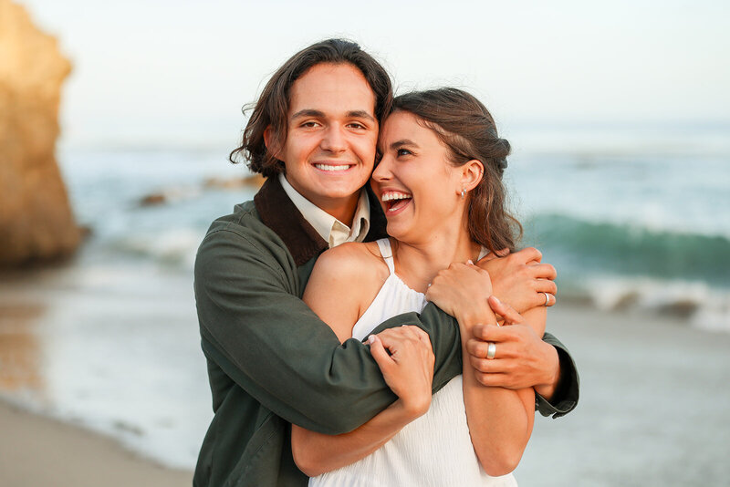 Engaged couple standing next to the ocean hugging