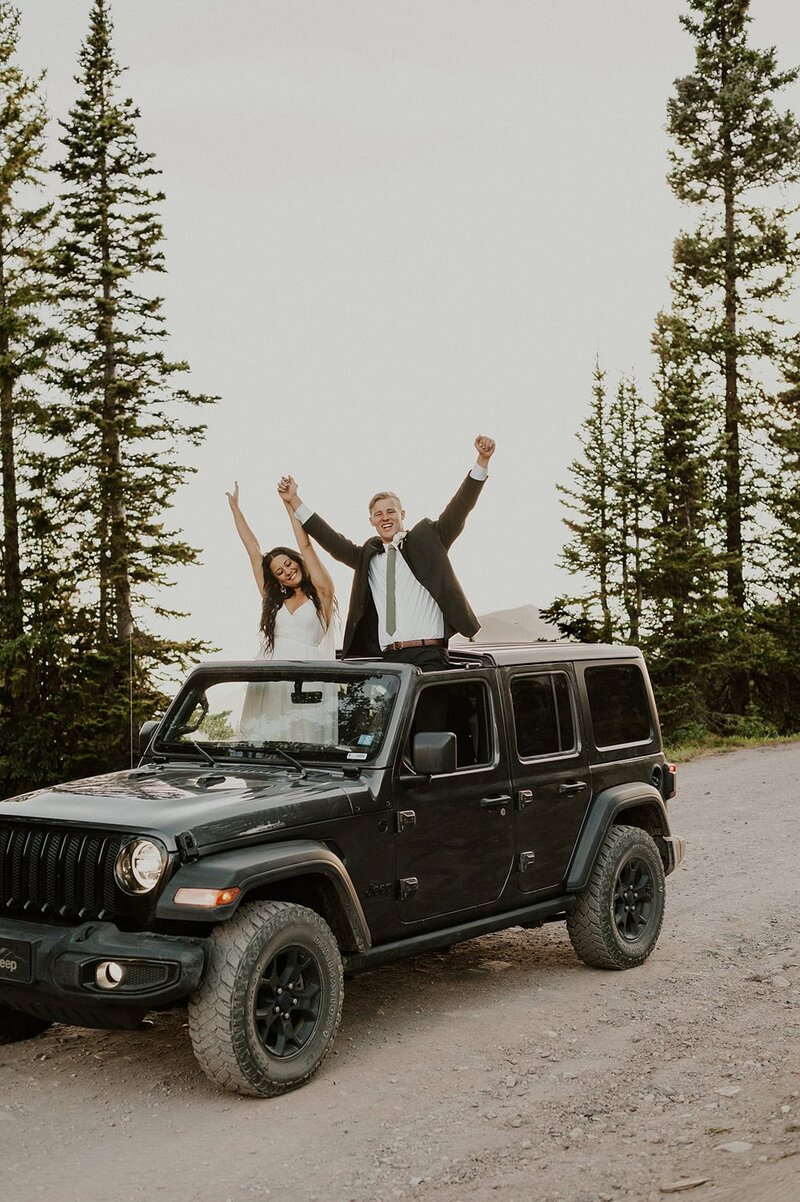 Couple in wedding attire cheering out of the top of their jeep, after their off-roading Colorado elopement in Crested Butte