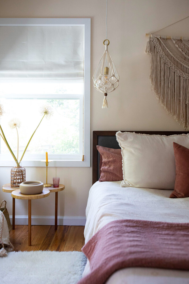 Bedroom featuring an art deco boho interior design. Brightly lit bedroom with a bed, macrame wall hanging a lamp, and a side table with various objects on it.