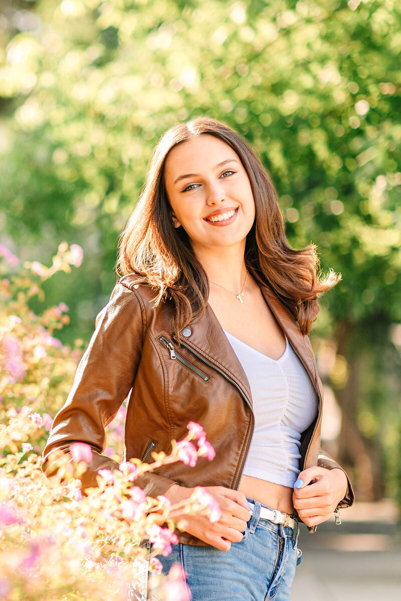High school senior girl wearing a brown leather jacket and white t-shirt in front of a bush with pink flowers for her Pleasanton CA senior photos