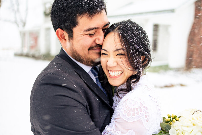 A wedding couple embracing in the falling snow and laughing after their wedding at Cornman Farms
