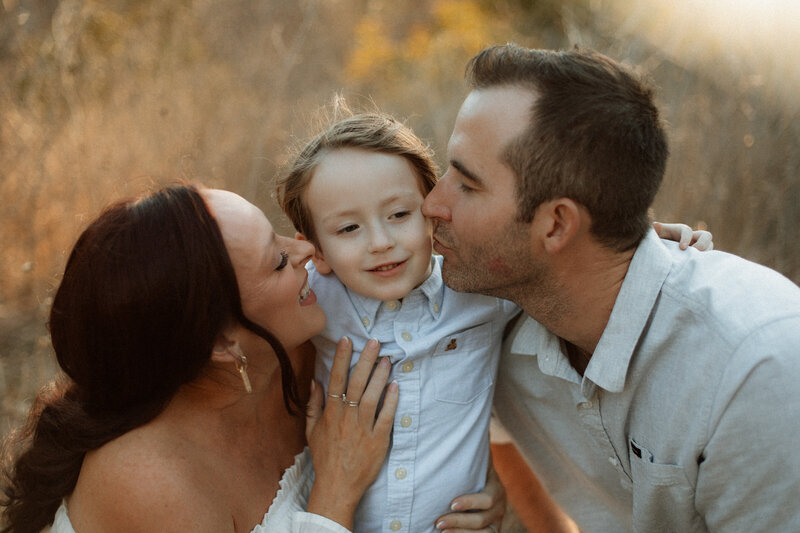 Smiling parents kissing their toddler's cheeks in soft golden light during a family session in Temecula.