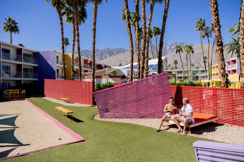 A stylish couple enjoys a laid-back moment at a vibrant Palm Springs resort, surrounded by colorful mid-century architecture and swaying palm trees. With desert mountains in the background and a playful, retro aesthetic, this image perfectly captures the essence of a relaxed and romantic getaway.