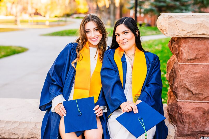 two NAU senior graduates  sitting on ledge on campus northern Arizona university in graduation attire holding caps