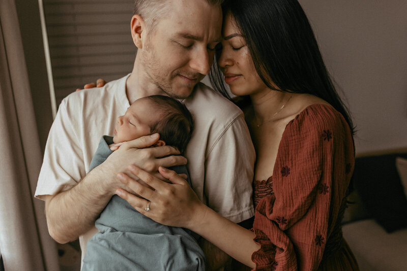 A mother and father hold each other while holding their newborn baby in their arms. 