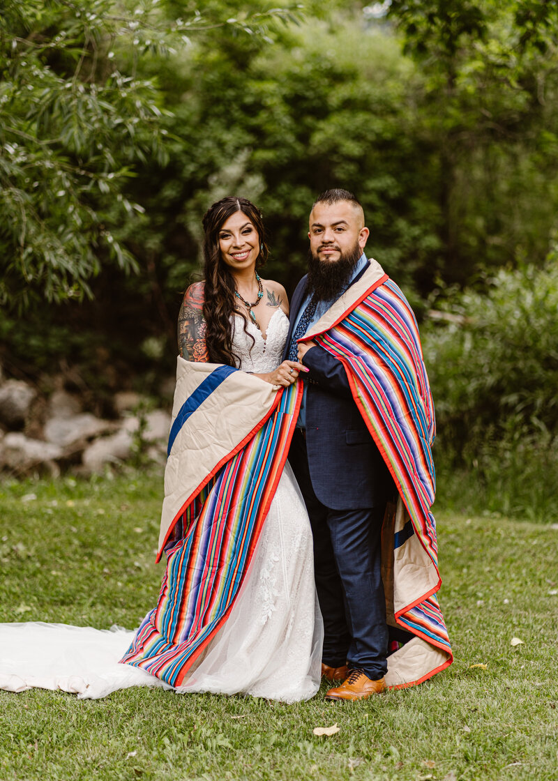 Couple stands side by side wrapped in a traditional Indigenous blanket after their wedding ceremony with Fort Collins, Colorado portrait photographer Avenir Photo Co.
