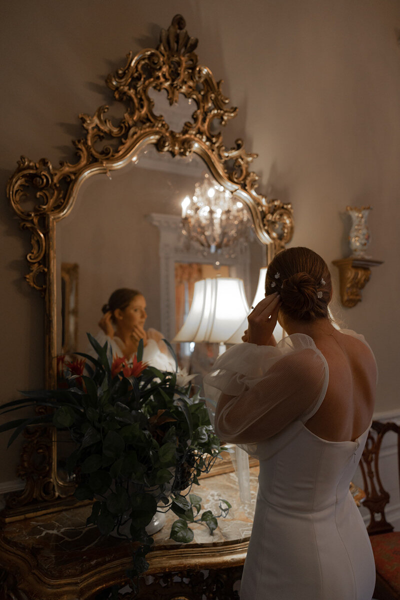Bride is looking at antique mirror  with golden frame and is fixing her bridal hair and does a final bridal makeup check befire getting bridal portraits taken.