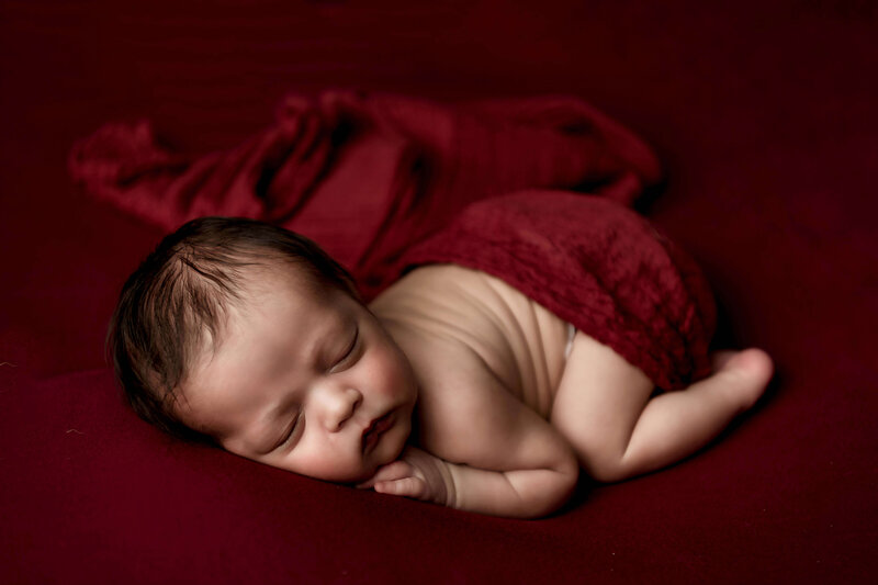 A squishy newborn sleeping peacefully in what is called the bum up pose. You can see his tiny newborn details perfectly, including the fat rolls on his back.  The lighting highlights his features perfectly, as the red backdrop and blanket compliment his gorgeous skin tone.