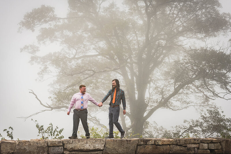 A gay couple walk on a stone wall hand-in-hand in front of a patch of fog covering a large tree at Loft Mountain Overlook in Shenandoah National Park.