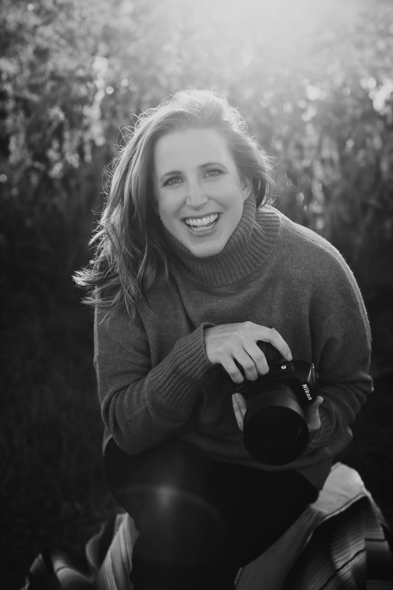 Black and white image of Nicole Rose backlit sitting in a field wearing a turtle neck sweater and dark pants holding a digital camera