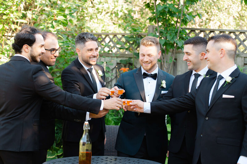 a groom and his groomsmen in black tuxedoes toasting on wedding day.  Captured outdoors by Ottawa wedding photographer JEMMAN Photography