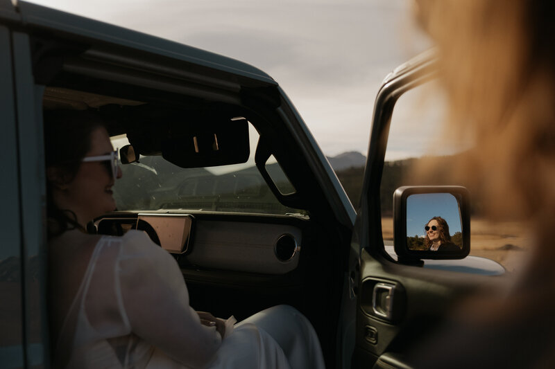 A newlywed sitting in a Jeep as their partner's standing next to them with their reflection in the side mirror 