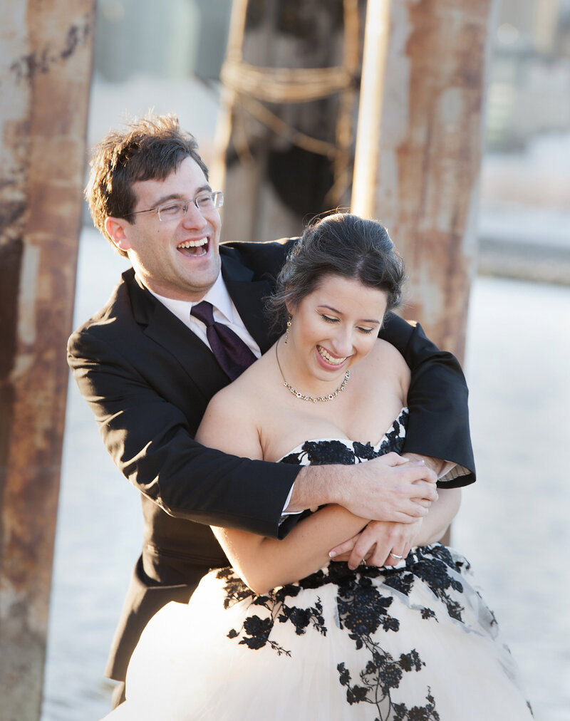 A person standing behind their partner with their arms wrapped around them as they both laugh 