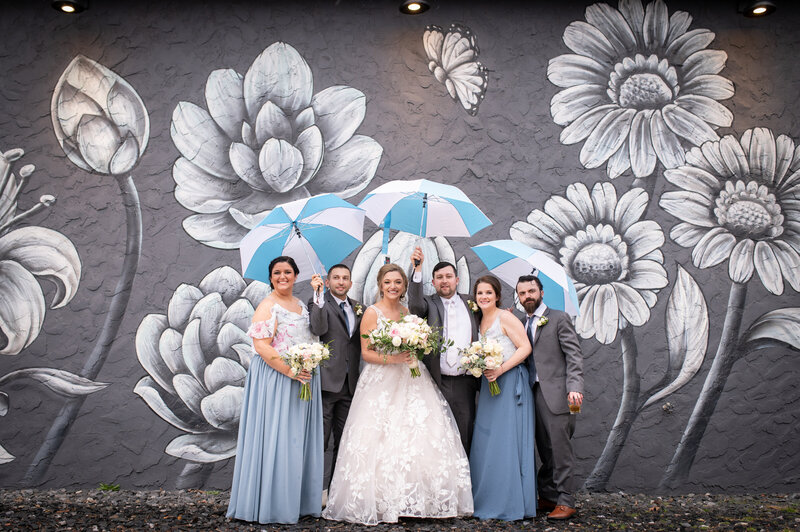 bridal party with bride and groom huddle under blue and white umbrellas in front of a a black and white floral mural