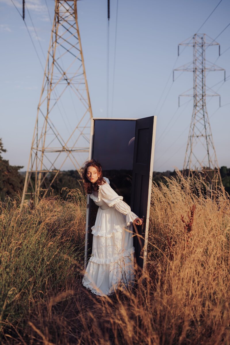 Woman wearing a flowy white dress in a field emerges through through a white door with powerlines behind her.