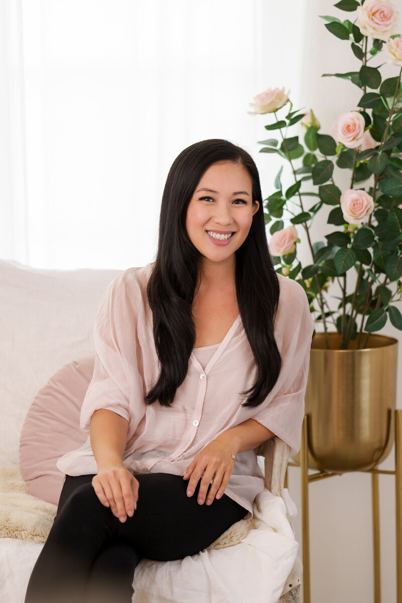 Asian woman with long dark brown hair in a light pink sheer top with a matching light pink tank top underneath and black pants sitting on a chair covered by a white blanket with a pink rose tree in a gold midcentury style tall plant holder.