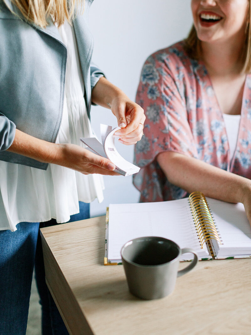 two people working together, one sitting at a desk with a notebook and coffee, the other standing with a notepad
