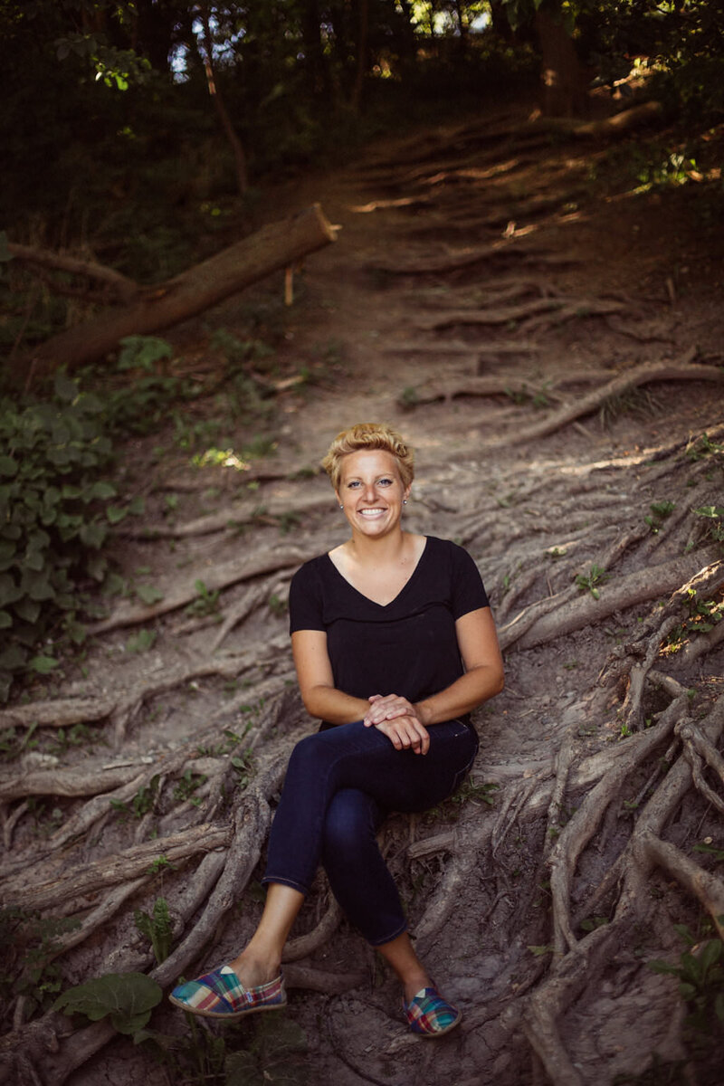 Writer, Sarah Freymuth, smiles for photo in black v-neck t-shirt while sitting on tree roots outdoors