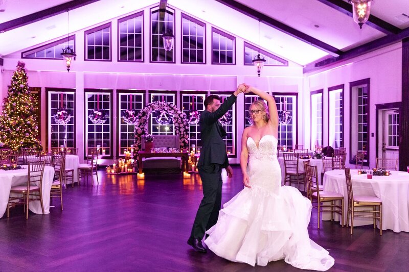 The bride, in a strapless mermaid-style wedding gown, twirls gracefully under the groom’s hand on the dance floor of their Pennsylvania wedding reception. The venue glows with soft purple uplighting, candles flickering beside a floral arch, and a towering Christmas tree adorned with twinkling lights