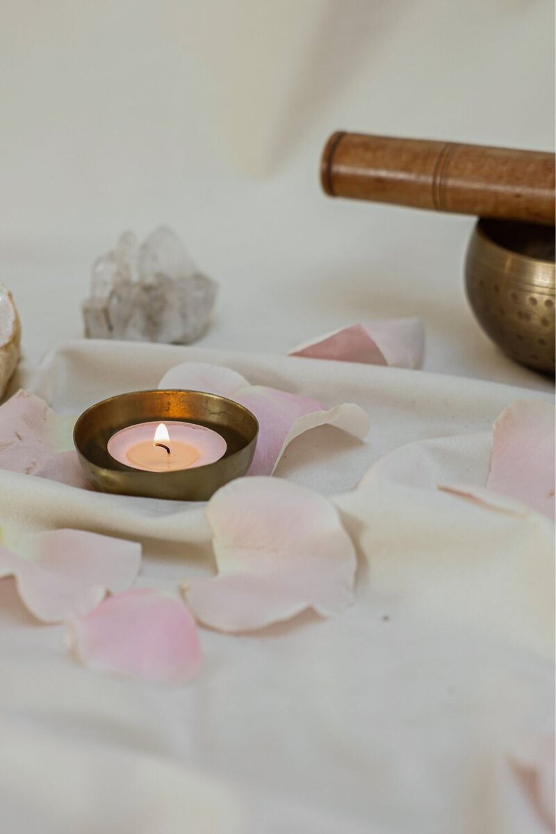 Lit candle in a brass holder surrounded by rose petals, crystals, and a sound bowl, evoking peace and energy healing