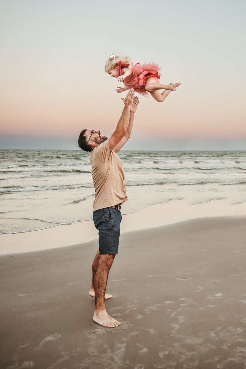 DAD THROWS TODDLER IN THE AIR WHILE SHE GIGGLES AT SUNSET AT PORT ARANSAS BEACH IN TEXAS