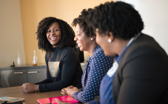 Three women smiling and conversing at a work table, representing potential university or corporate partners collaborating with the Center for Leadership Excellence in leadership training programs.