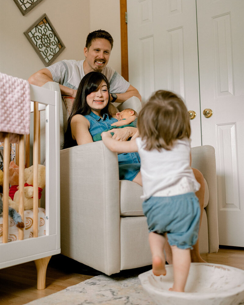 A toddler runs towards her mom and dad who are sitting in a chair with an infant