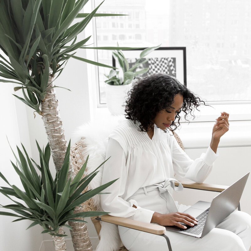 Woman with curly hair in a white outfit working on a laptop in a bright home office