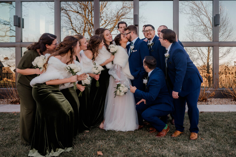 Bridal party huddles together smiling while watching the bride and groom share a kiss.