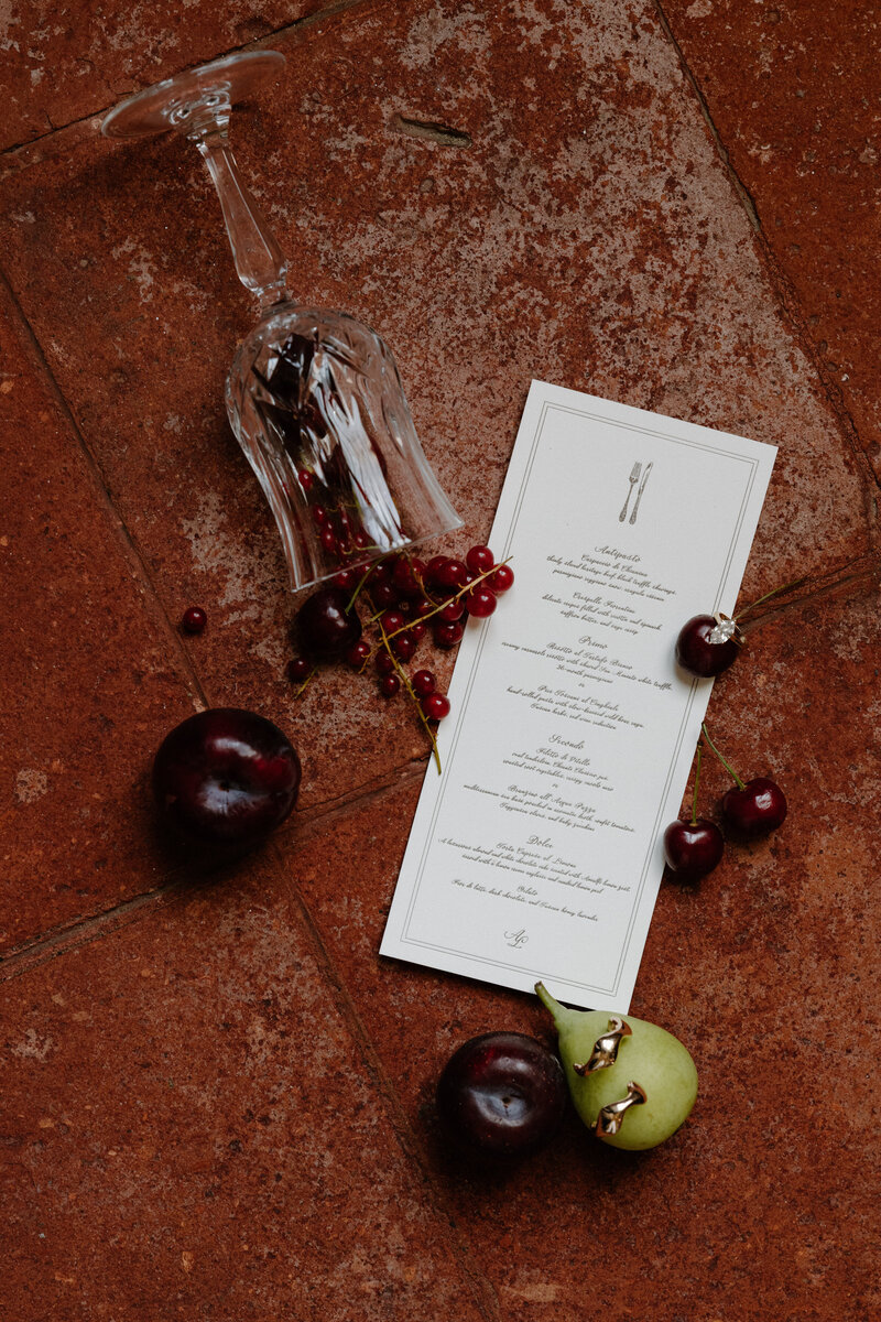 A glass overturned with fruit running out of it with a wedding invitation next to it and a small fruit with wedding rings inside of it 