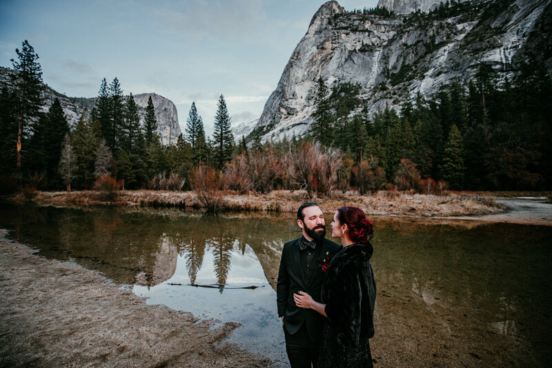 Moody winter elopement at Mirror Lake in Yosemite National Park, with a couple dressed in dark, romantic attire standing near still water reflecting towering granite cliffs and pine trees.
