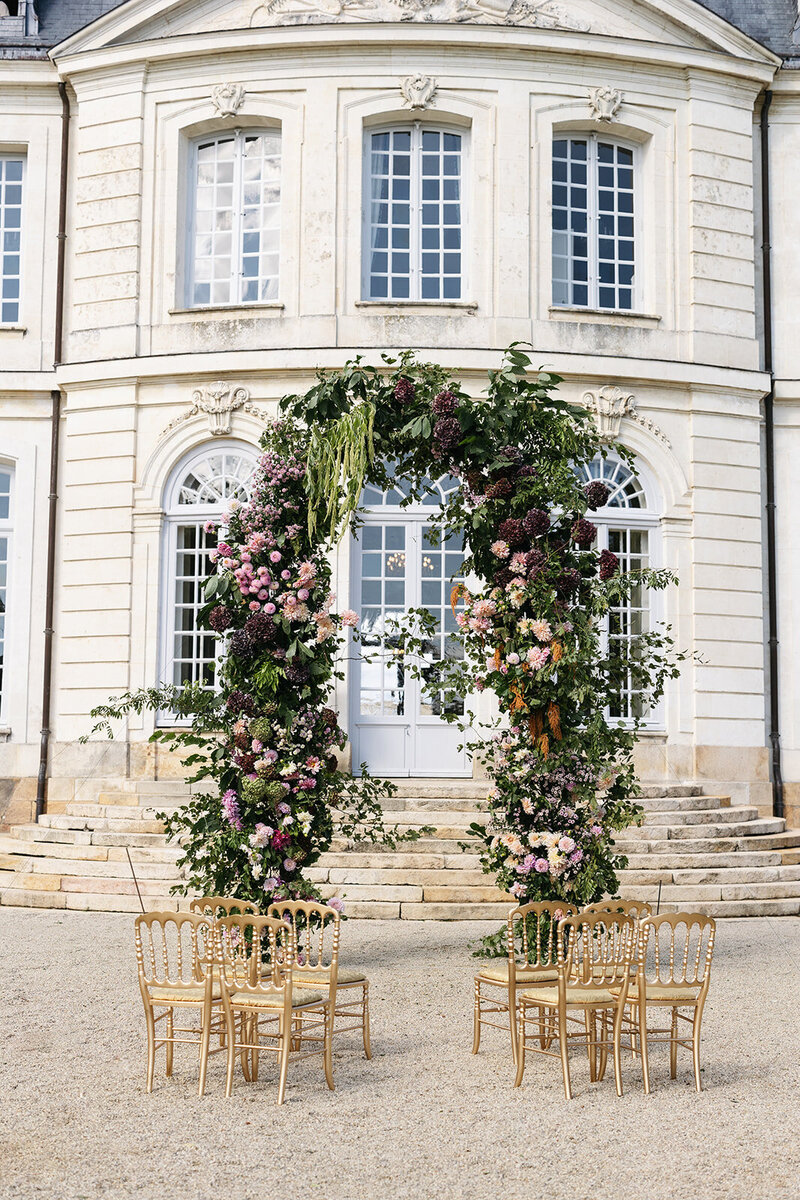 A grand floral arch featuring lush greenery and colorful blooms stands in front of a stately French chateau with elegant windows.