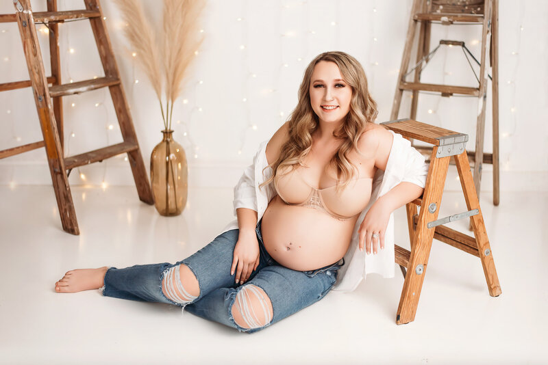 A pregnant woman in jeans and a white button up leaning on a ladder for boho maternity photos in a downtown studio