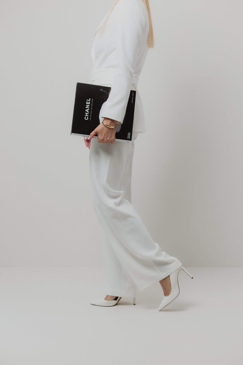 A women in a white power suite and heels walking and holding a Chanel book.