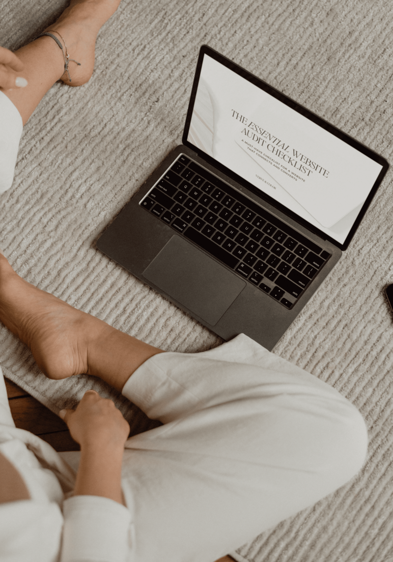 Vickie sitting on a cozy rug with a laptop displaying ‘The Essential Website Audit Checklist.
