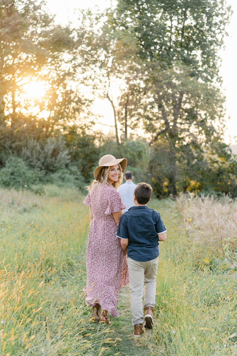 Mom in maxi dress and hat walks through a field of golden grass with her teen son