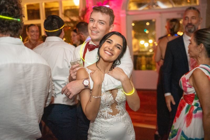 A joyful bride in a white lace wedding dress and groom in white shirt with red bow tie embrace while dancing at their wedding reception at the White Room in historic downtown St. Augustine, surrounded by guests and colorful lighting