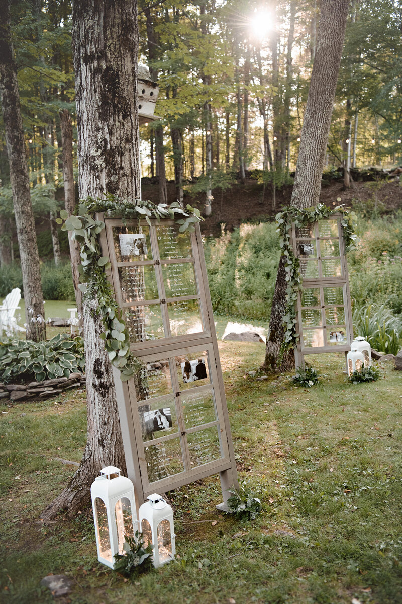A rustic modern seating chart made from reclaimed windows and surrounded by white lanterns leans against a tree beside a pond at a Catskills wedding venue