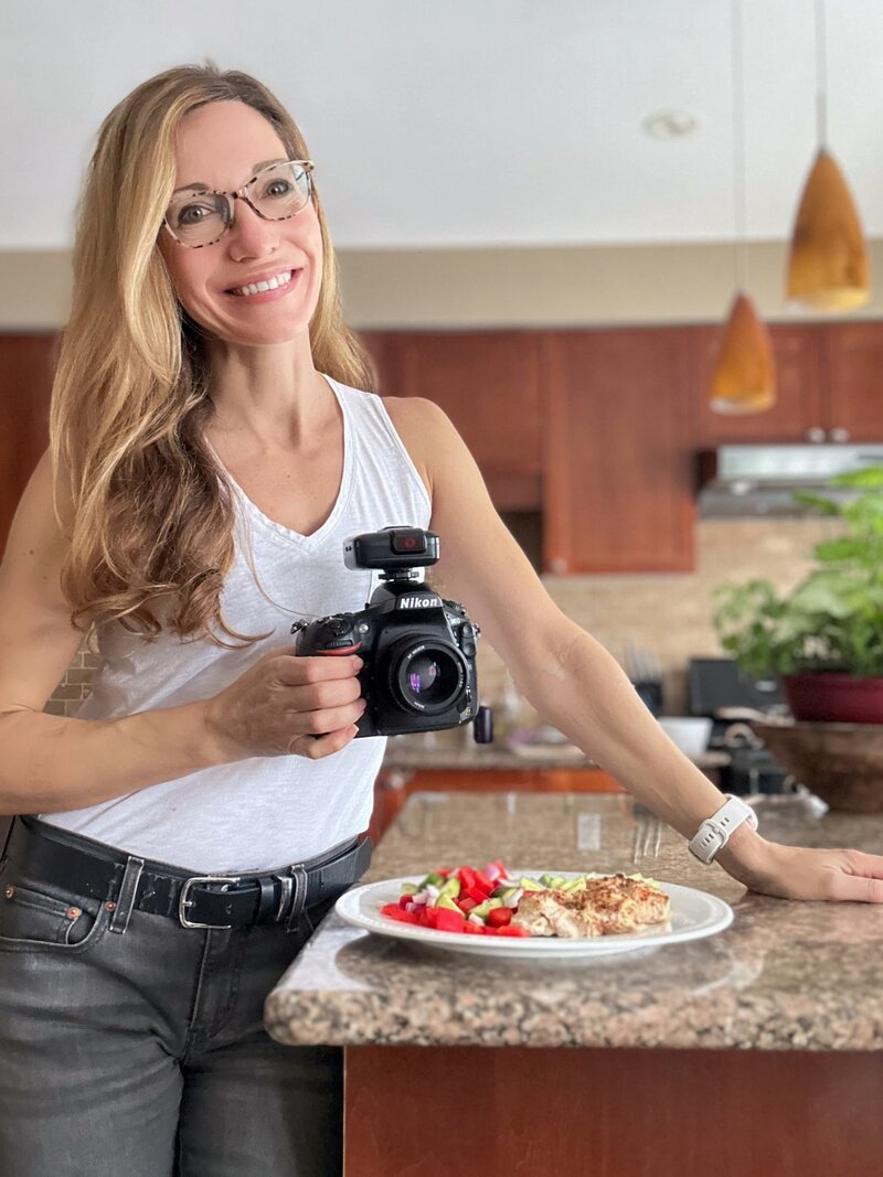 Toronto Photographer Carla Morgado standing in Kitchen photographing food and smiling at the camera