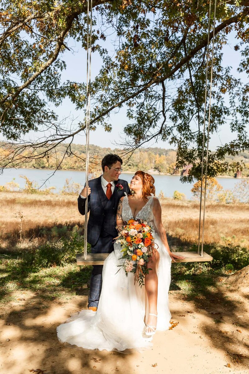 A newlywed couple poses on a wooden swing under a sprawling tree at Tennessee River Place, with a scenic view of the Tennessee River in the background. The bride, in a lace gown with a thigh slit, holds a vibrant bouquet of orange, peach, and pink flowers, gazing at her groom, who wears a navy plaid suit with a burnt orange tie