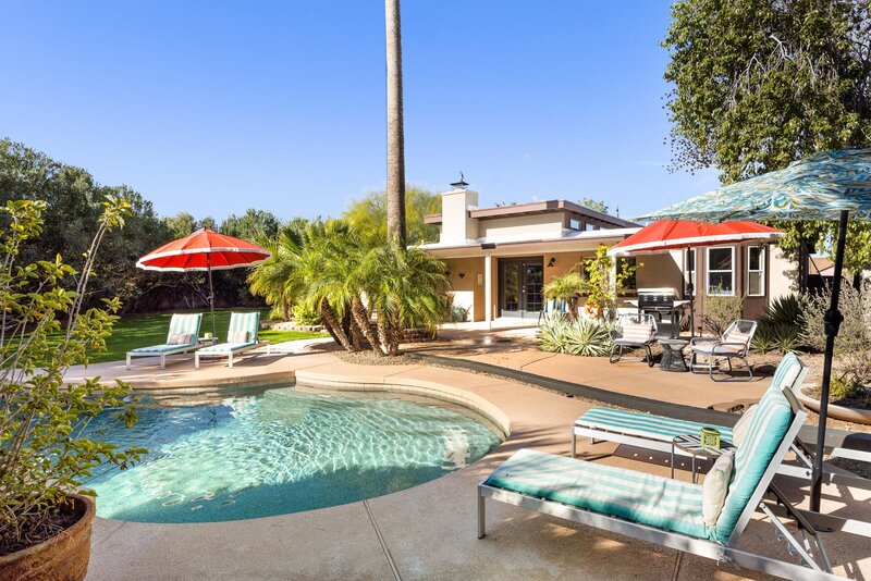 Backyard in Arizona with a sparkling blue pool and lush palm trees. There are lounge chairs and orange umbrellas with fringe.
