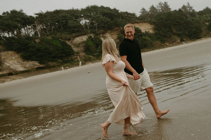 Couple skipping along the beach at Seal Rock, Newport, Oregon, with the coastal waves and dramatic rock formations in the background.