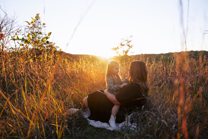 Daughter sitting in mother's lap in a field at sunset with the glowing sun behind them in De Soto, Missouri. 