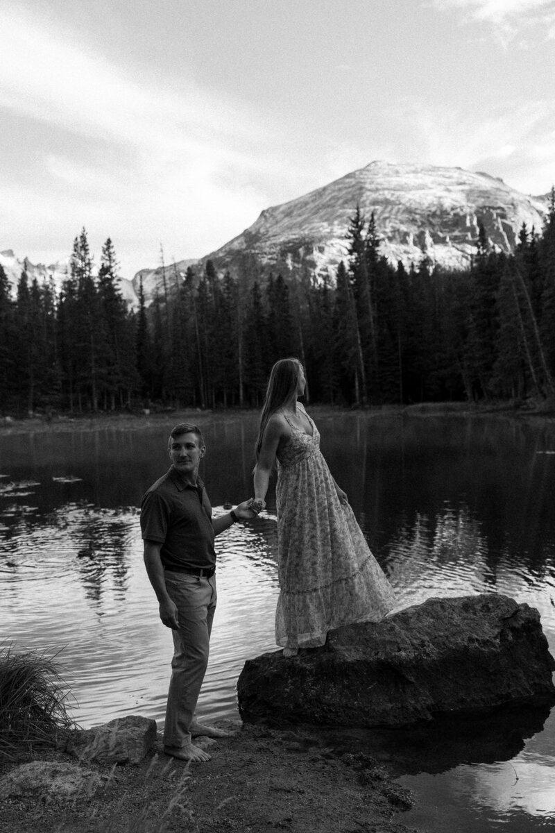 Man and woman holding hands while overlooking a lake with mountains in the background, captured by Andrea-Marie Photography
