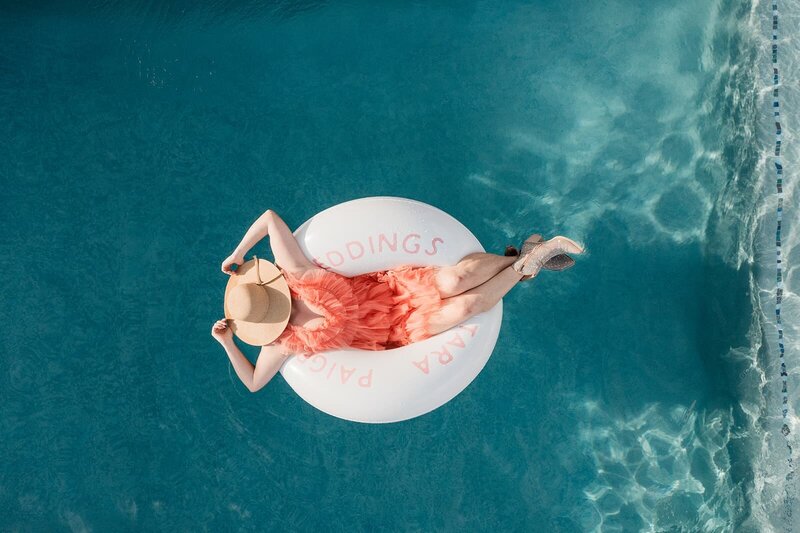 Overhead view of a woman in a straw hat and coral dress floating on a custom pool float labeled “Tara Paige Weddings” in a serene blue pool.