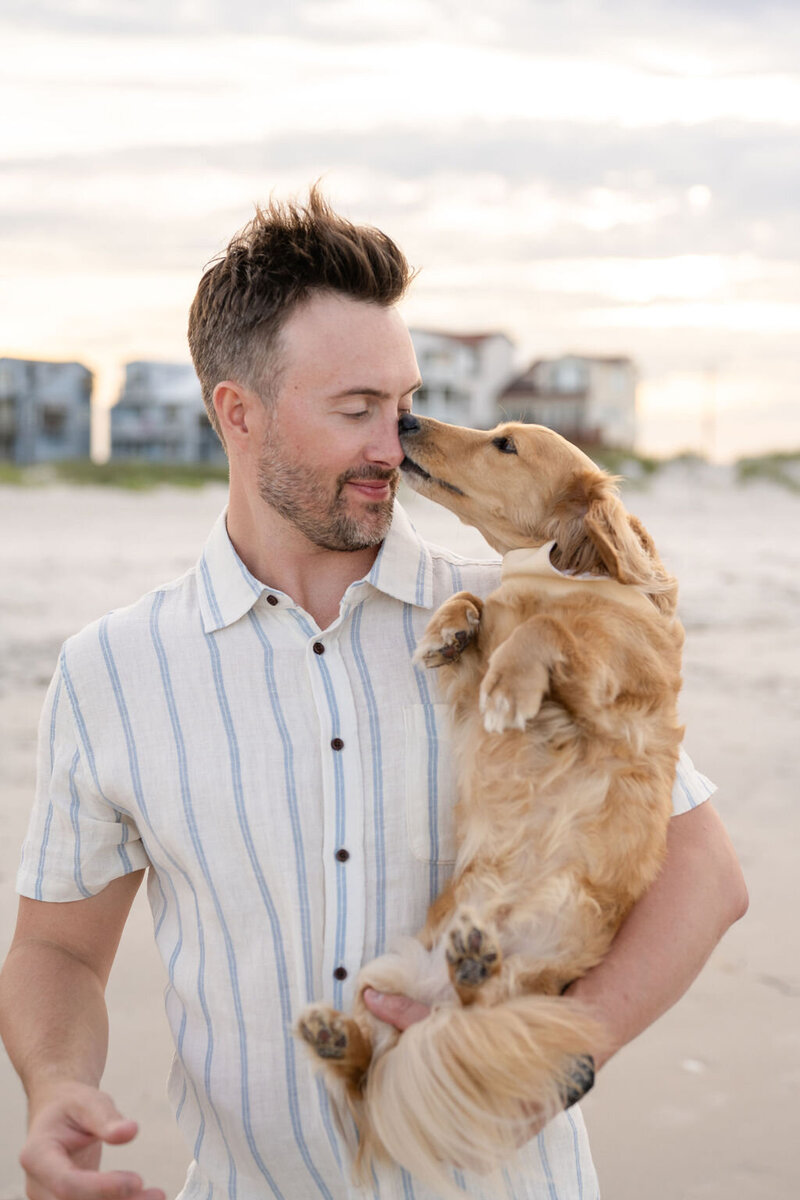 man stands on the beach cradling his cute dog as it snuggles up to him