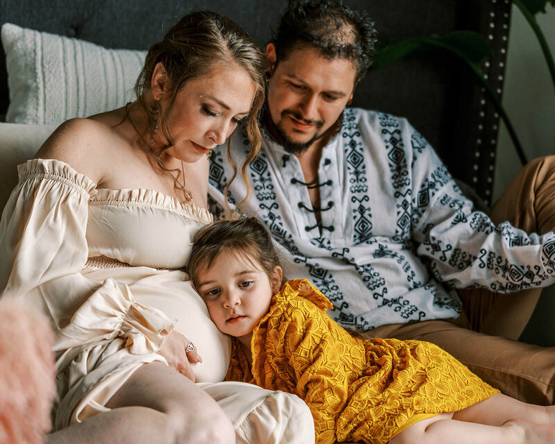A child snuggles with her mom and dad and rests her head against her mom's pregnant belly. 