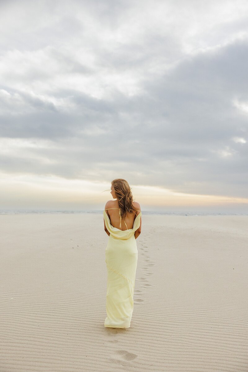 Editorial senior portrait of girl in yellow gown walking across the sand at a Santa Monica beach in Los Angeles at sunset